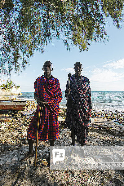 Zwei Massai-Männer in traditioneller Kleidung stehen bei Sonnenuntergang am Strand