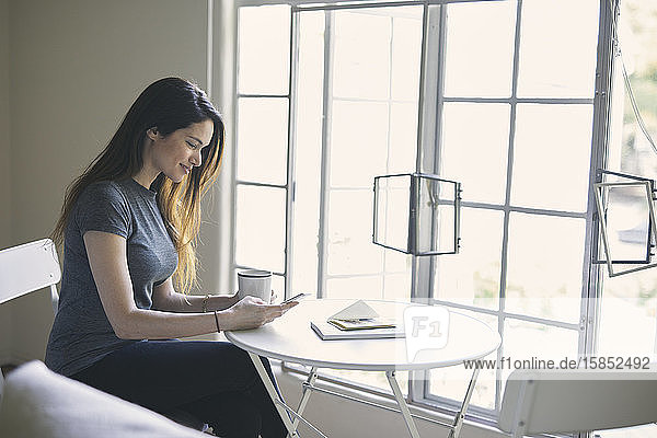 Junge Frau sendet Textnachrichten auf einem Smartphone  während sie im Wohnzimmer Kaffee am Tisch trinkt