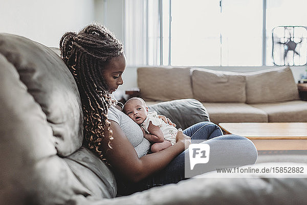 Ethnic mother with long braids on sofa holding multiracial newborn