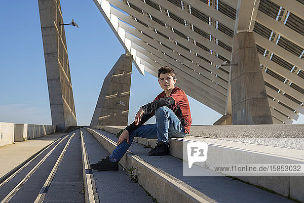 Junger Teenager sitzt auf einer Treppe im Freien und schaut bei Sonnenschein in die Kamera