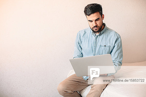 Junger Mann benutzt Laptop-Computer  während er zu Hause an der Wand sitzt