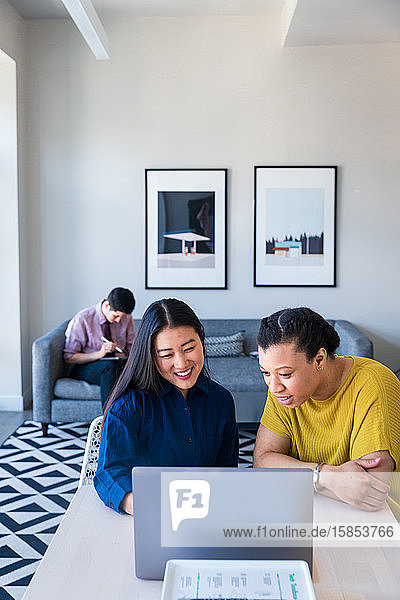 Lächelnde Unternehmerinnen planen Strategie über Laptop  während ein männlicher Kollege im Büro auf dem Sofa sitzt