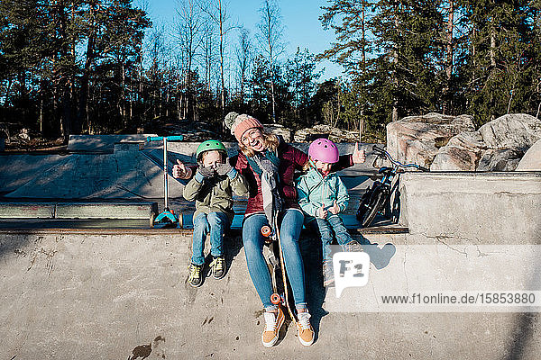 Mama und ihre Kinder spielen in einem Skatepark mit Rollern und Fahrrädern