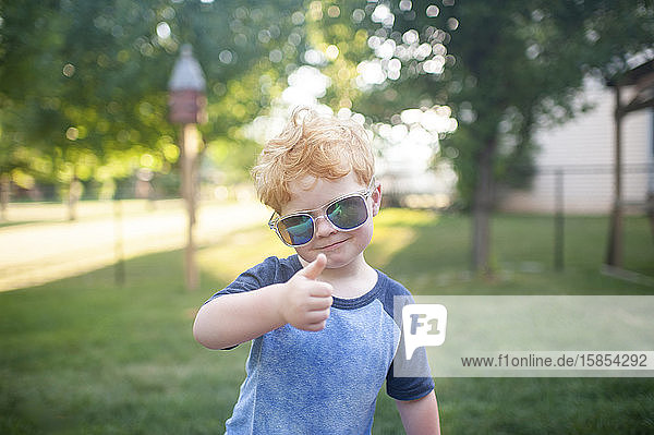Young boy giving a thumbs up with sunglasses on standing in backyard