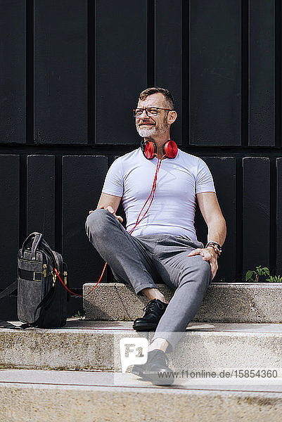 Bärtiger  reifer Mann sitzt mit Kopfhörern auf Stufen und entspannt sich an einem sonnigen Tag