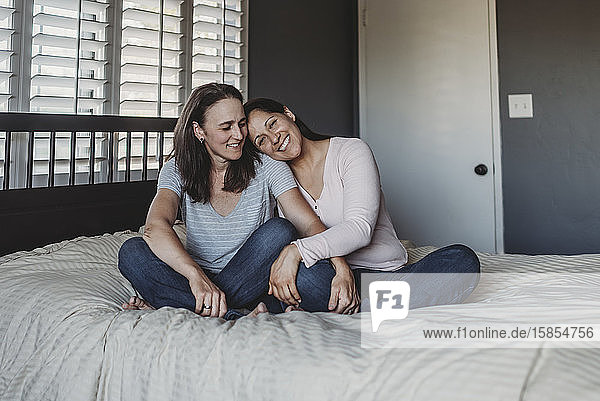 Lächelndes gleichgeschlechtliches Paar kuschelt sich auf einem Bett in Fensternähe mit Fensterläden