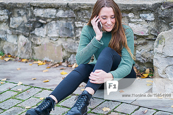 portrait of a young Caucasian woman with brown hair sitting with her back supported by a stone bridge and talking on her mobile phone. Dressed in a green sweater  dark pants and military boot