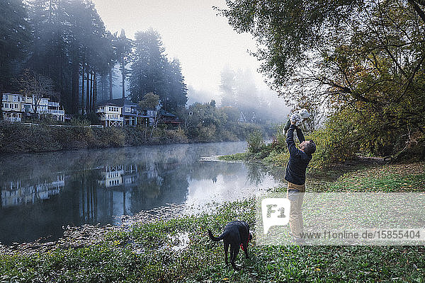 A man is holding a baby near a river and a dog