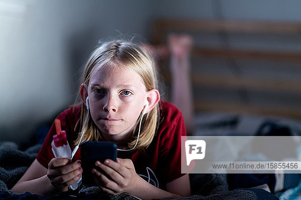Teenager hält Eis am Stiel mit Telefon und Ohrstöpseln auf dem Bett liegend