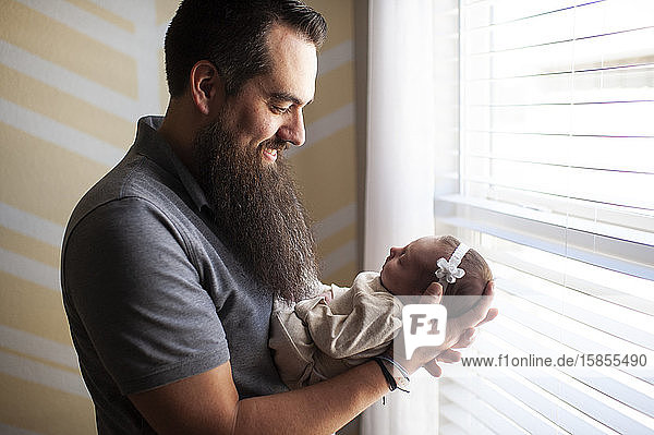 Vater lächelt  während er seine neugeborene Tochter zu Hause am Fenster hält