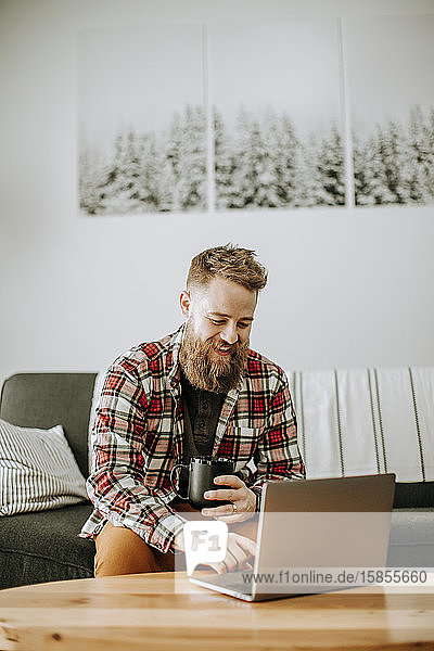 man with beard holds cup of coffee while working on laptop computer