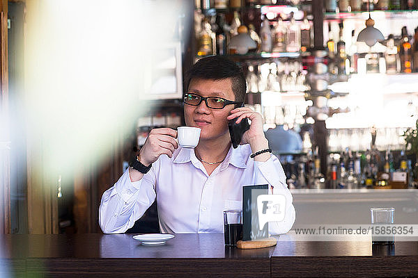 junger asiatischer Mann in einer Bar mit einer Tasse Kaffee und einem Smartphone