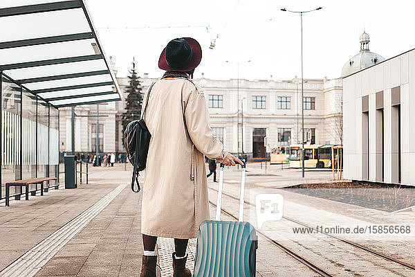 Stilvolles Mädchen mit einem Koffer steht an einer Stadthaltestelle und wartet auf eine Straßenbahn