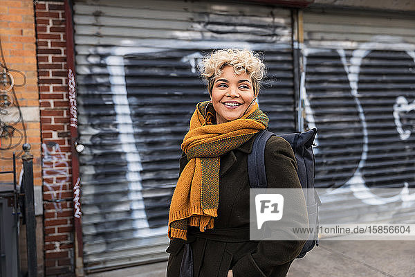 Lächelnde Frau mit Schal schaut weg  während sie in der Stadt gegen einen geschlossenen Fensterladen steht