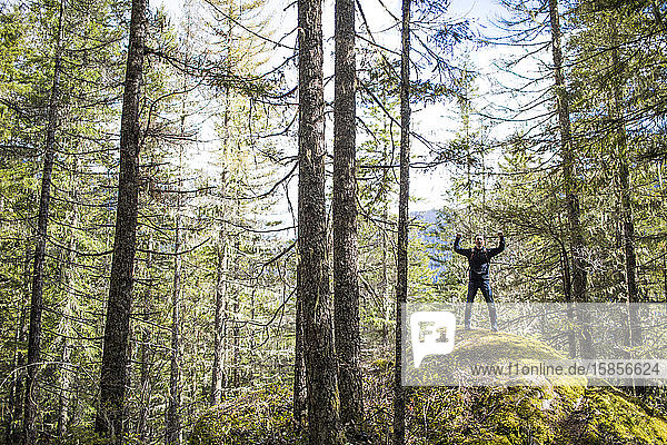 Wanderer hebt die Arme  nachdem er einen moosbedeckten Felsbrocken im Wald erobert hat.