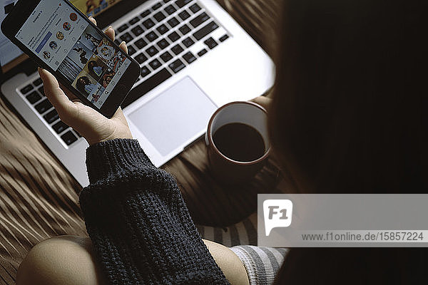 VSETIN  CZECH REPUBLIC/28 October 2019: Young girl in winter socks sitting on the bed with laptop  holding cup of coffee and watching instagram on her smartphone. Winter  cozy  clothes and li