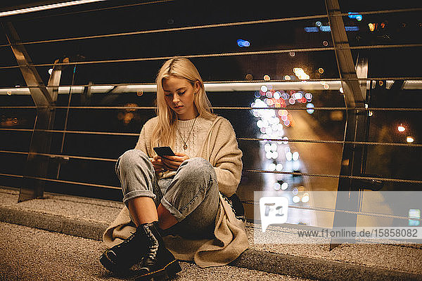 Teenager-Mädchen benutzt Smartphone und sitzt nachts auf einer Brücke in der Stadt