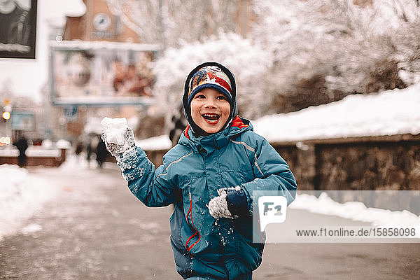 Happy boy holding snowball while running in street during winter
