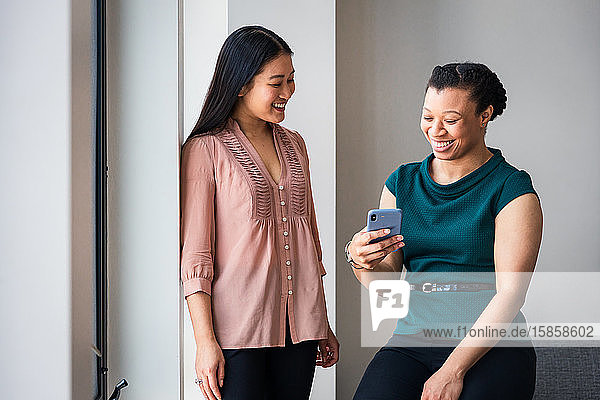 Happy female colleagues looking at smart phone against wall in office