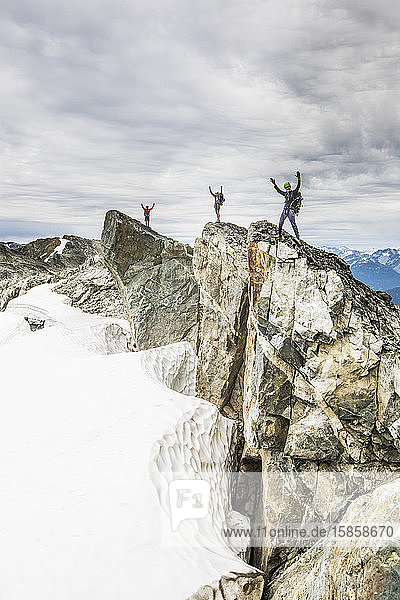 Three climbers stand on a rocky summit with arms raised in the air