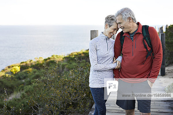 Happy senior couple with arm in arm standing on boardwalk by sea against sky