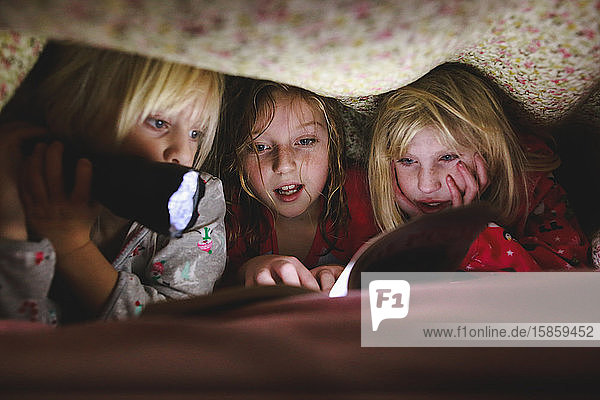 Three little girls reading book under blanket with flashlight