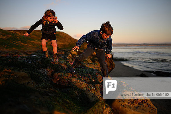 Siblings climbing down rocks near the ocean at low tide and sunset