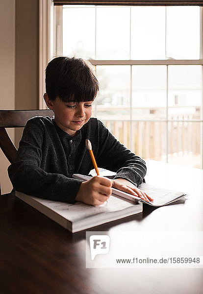 Young boy doing schoolwork in a workbook at home at the table.