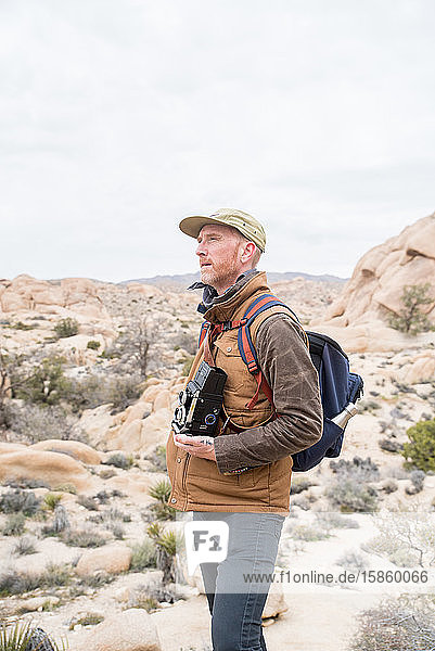 Man with billed hat and camera in desert by stones and plant life