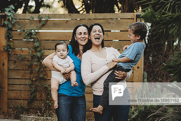 Laughing moms holding young boy and baby in backyard with wooden fence