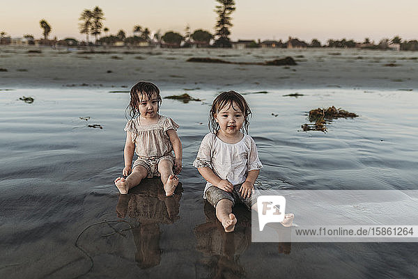 Front view of toddler sisters sitting in water at beach