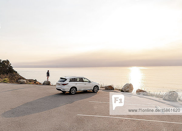Ein am Aussichtspunkt Big Sur geparktes Auto mit einer auf einem Felsen stehenden Frau