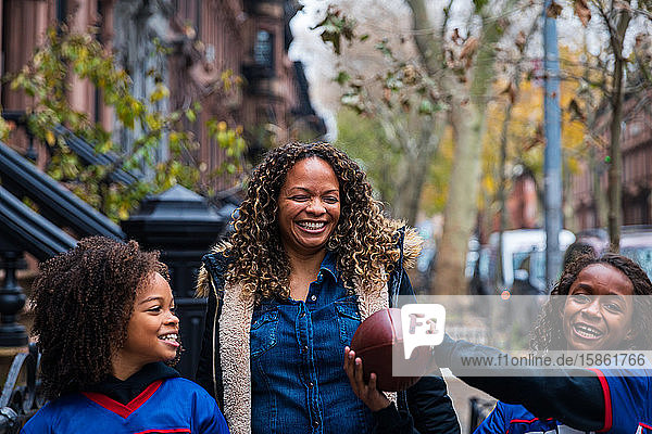 Glückliches Mädchen hält American Football in der Hand  während es Schwester und Mutter auf einem Fußweg in der Stadt zur Seite steht