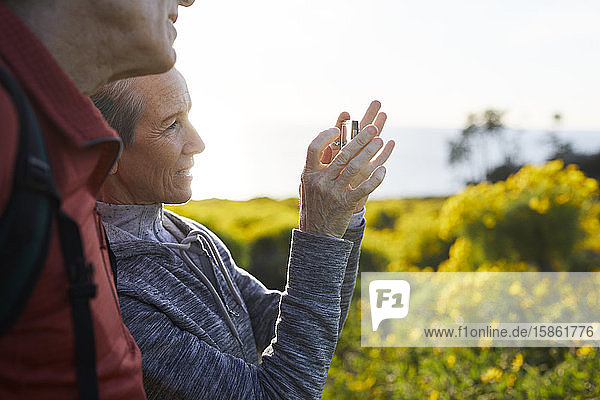 Mann steht neben älterer Frau  die mit einem Smartphone fotografiert  während sie sich gegen den Himmel stellt