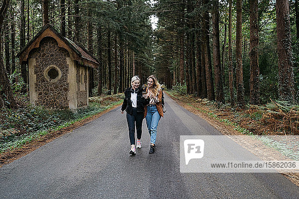 Two women walking on a road in the forest while watching their phone