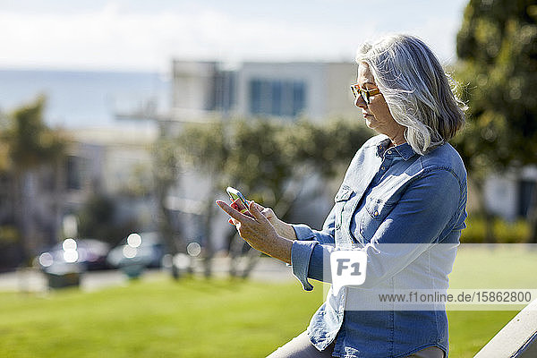 Frau trägt ein Jeanshemd und benutzt ein Smartphone  während sie an einem sonnigen Tag im Park steht