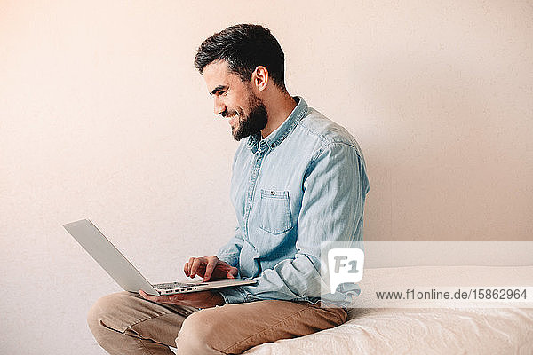 Glücklicher Mann benutzt Laptop-Computer  während er zu Hause an der Wand sitzt