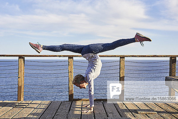 Full length senior woman doing handstand while exercising on floorboard by sea against sky