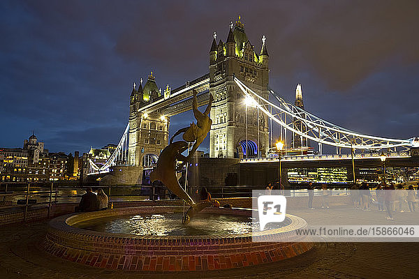 Tower Bridge on the River Thames and The Shard illuminated at dusk with a fountain and tourists in the foreground; London  England
