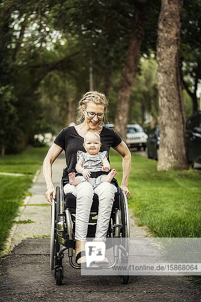 Eine querschnittsgelähmte Mutter  die ihr Baby auf dem Schoß trägt  während sie einen Rollstuhl benutzt  an einem warmen Sommernachmittag im Freien: Edmonton  Alberta  Kanada