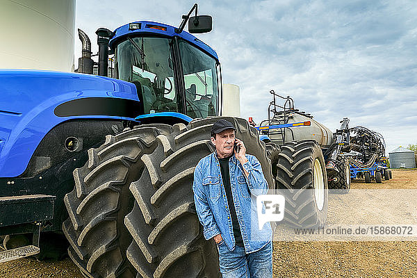 Landwirt  der ein Smartphone benutzt  während er auf einer Farm neben den Maschinen steht; Alberta  Kanada