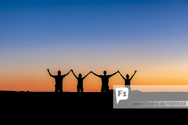 Silhouette von vier Menschen  die sich mit erhobenen Armen bei Sonnenuntergang in Aluvlei  Namib-Naukluft-Nationalpark  Namibia  an den Händen halten