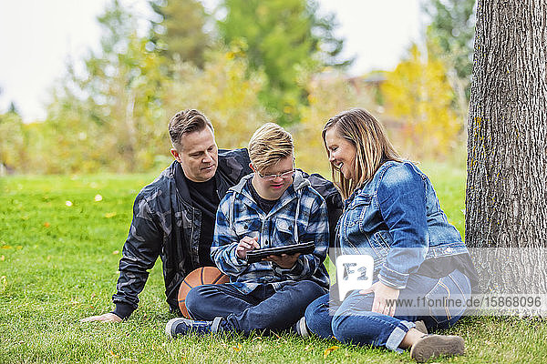 A young man with Down Syndrome learns a new program on a tablet with his father and mother while enjoying each other's company in a city park on a warm fall evening: Edmonton  Alberta  Canada