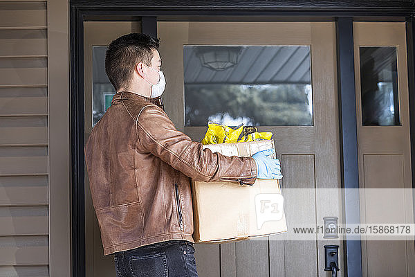 Ein junger Mann steht mit einer Kiste voller Lebensmittel vor der Tür eines Hauses während der Weltpandemie Covid-19; Edmonton  Alberta  Kanada
