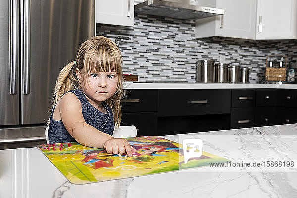 A cute little girl reading a book in the kitchen and looking up at the camera: Edmonton  Alberta  Canada