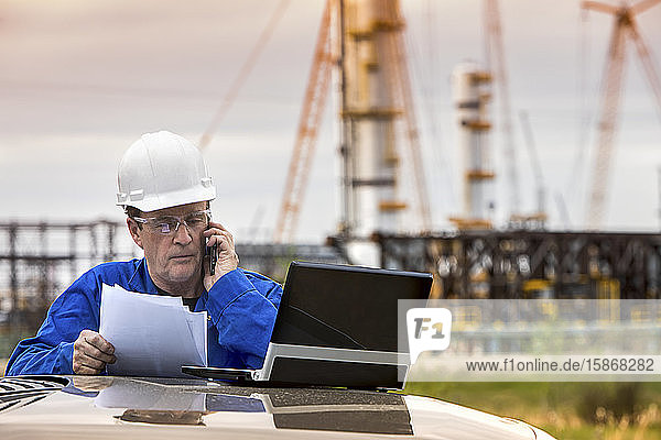 Man works on laptop and smart phone while standing at a vehicle with an oil rig in the background; Alberta Canada