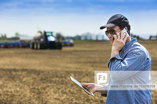 Landwirt  der ein Smartphone und ein Tablet benutzt  während er auf einem landwirtschaftlichen Feld steht und den Traktor und die Geräte beim Säen des Feldes beobachtet; Alberta  Kanada