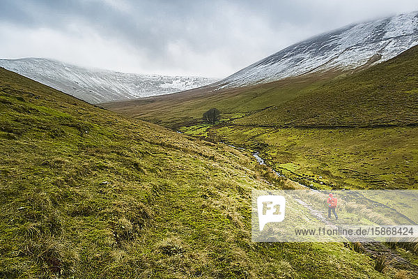 Lone female hiker in red raincoat carrying a red backpack walking along a valley trail leading to snow-covered mountains on a cloudy winter day  Galty Mountains; County Tipperary  Ireland