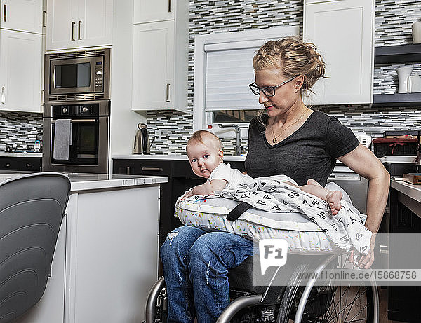 A paraplegic mother holding her baby on her lap  in her kitchen  while pushing in her wheel chair: Edmonton  Alberta  Canada