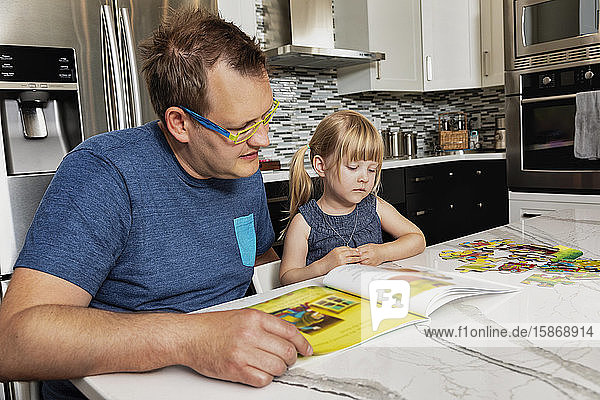 A father sitting down with his young daughter in the kitchen to read a book: Edmonton  Alberta  Canada